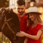 Charming young couple stands with a brown horse before a country house in the rays of evening sun