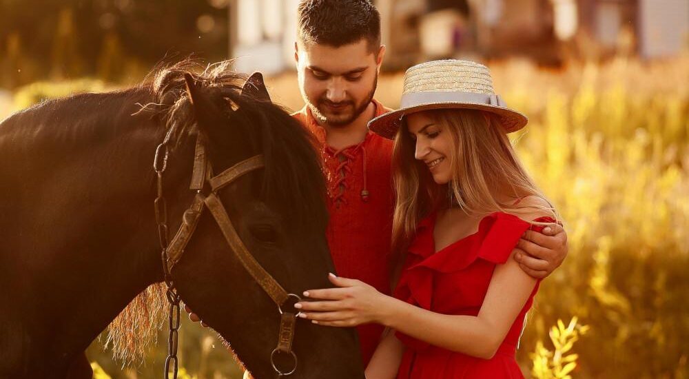 Charming young couple stands with a brown horse before a country house in the rays of evening sun