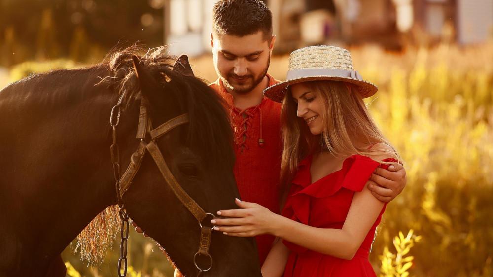 Charming young couple stands with a brown horse before a country house in the rays of evening sun